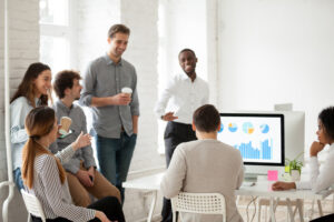 group of co-workers standing and sitting around a table, drinking coffee and talking.