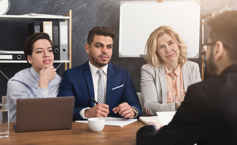 a group of people sitting around a table with 3 of them on one side talking to another on the other side.