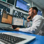 man sitting at desk surrounded by computers, keyboards, and monitors while on the phone.