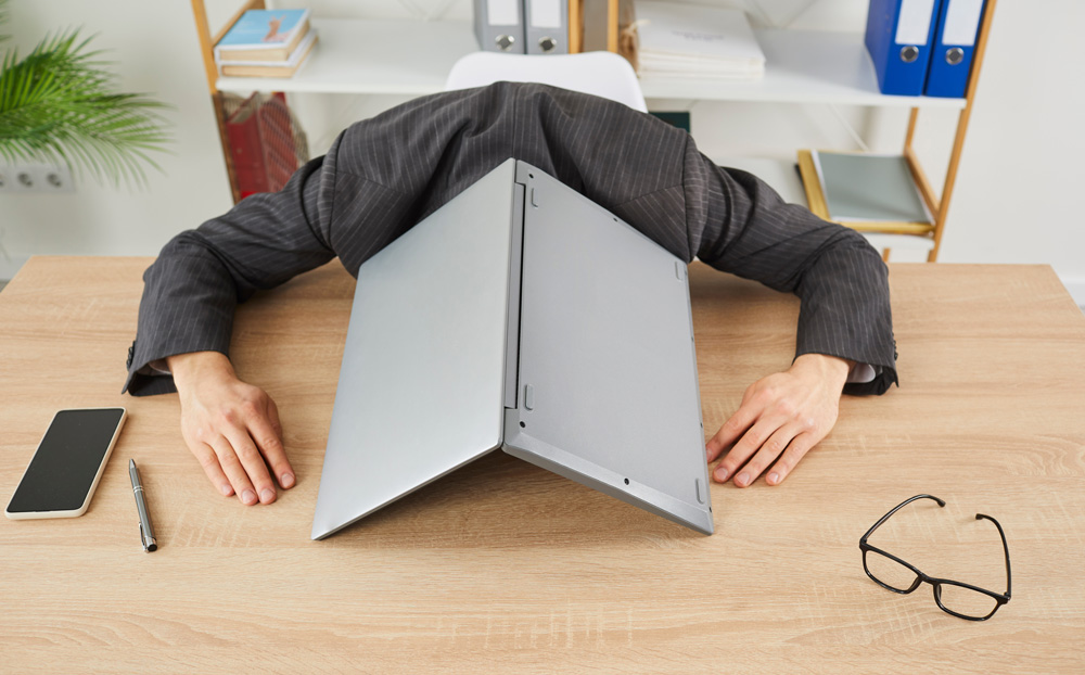 man in suit with head on table with laptop laying over his head